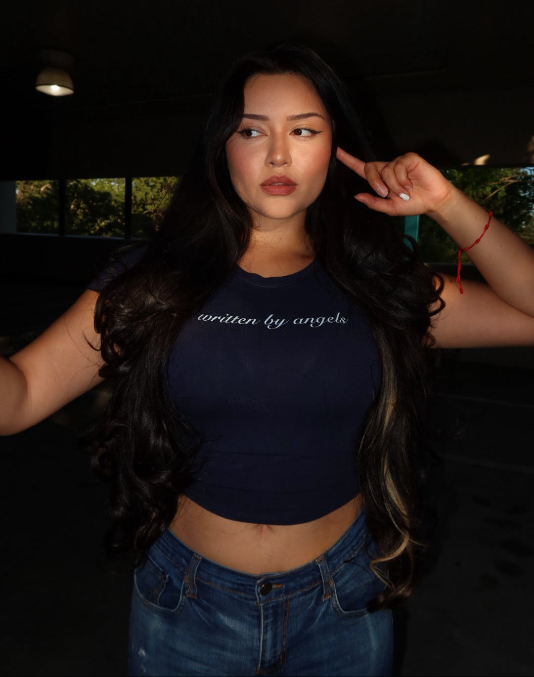 A woman with long, wavy dark hair wearing a navy blue crop top with 'written by angels' printed in white script across the chest, paired with blue jeans, posing in a dimly lit environment.